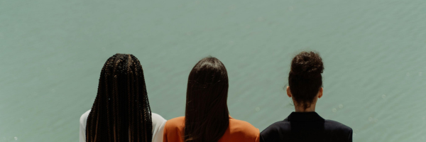 Three people are seen from behind, standing side by side and facing a calm body of water.