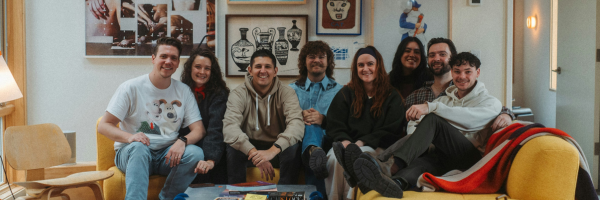 Group of seven people sitting closely on and around a yellow couch in a cozy, art-filled living room.